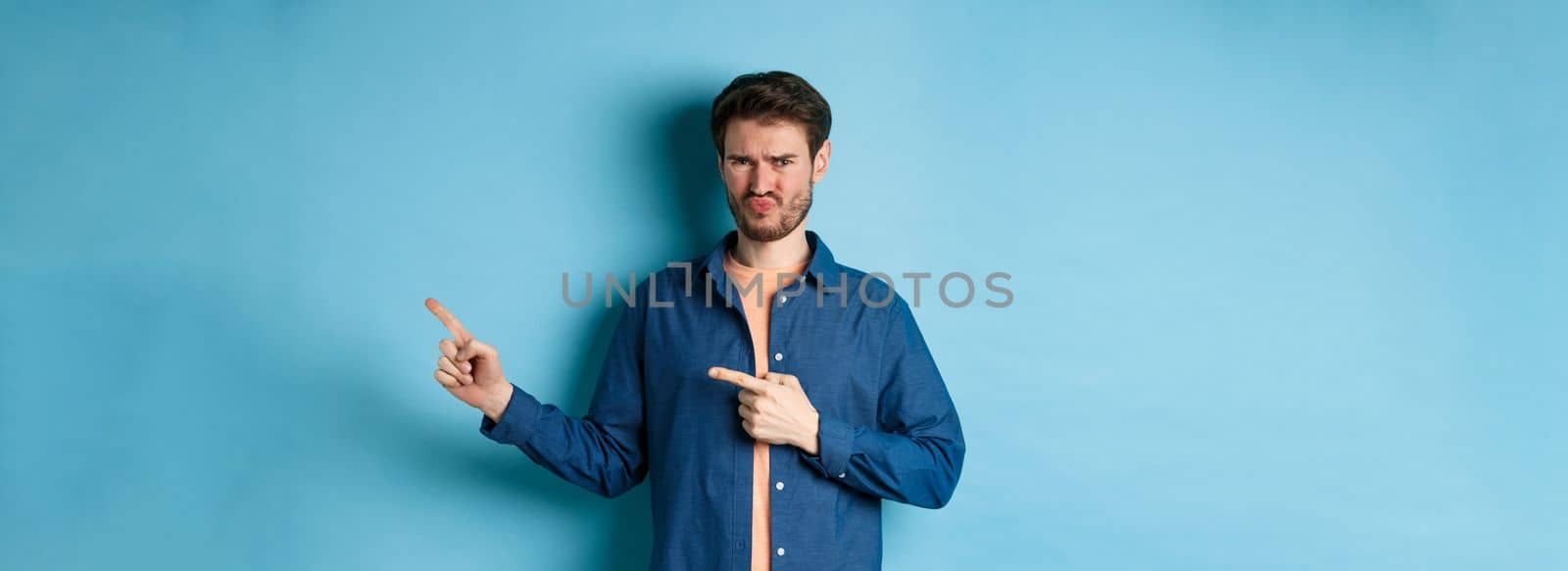 Disappointed young man frowning and pointing fingers right at empty space, looking upset and displeased, complaining, standing on blue background.