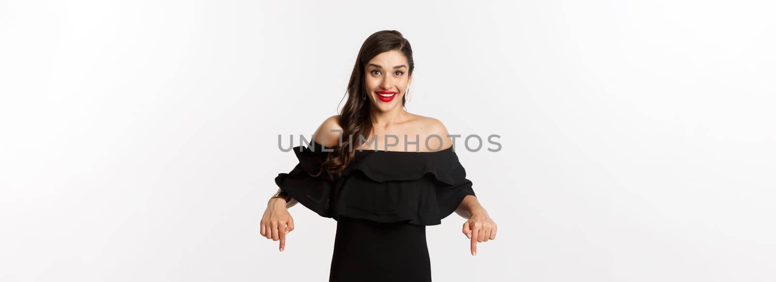 Fashion and beauty. Elegant woman in black dress pointing fingers down, showing promo and smiling, standing over white background.