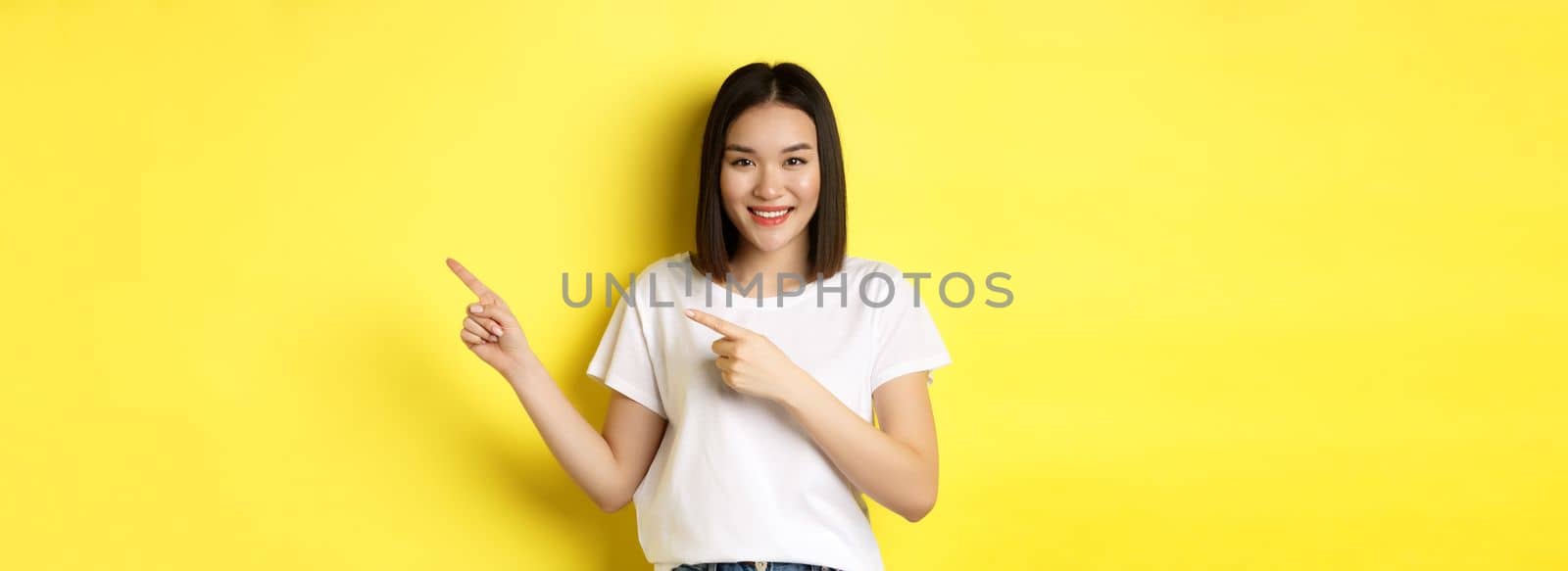 Beauty and fashion concept. Beautiful asian woman in white t-shirt pointing fingers right, demonstrate logo standing over yellow background.