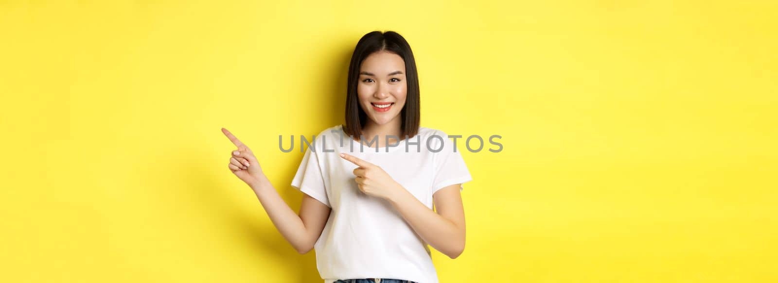 Beauty and fashion concept. Beautiful asian woman in white t-shirt pointing fingers right, demonstrate logo standing over yellow background by Benzoix