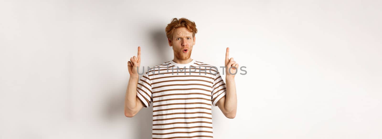 Confused young man with red curly hair pointing fingers up, staring shocked and puzzled, standing over white background.