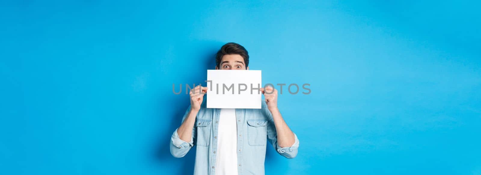 Excited man hiding face behind blank paper, place for your logo, standing over blue background.