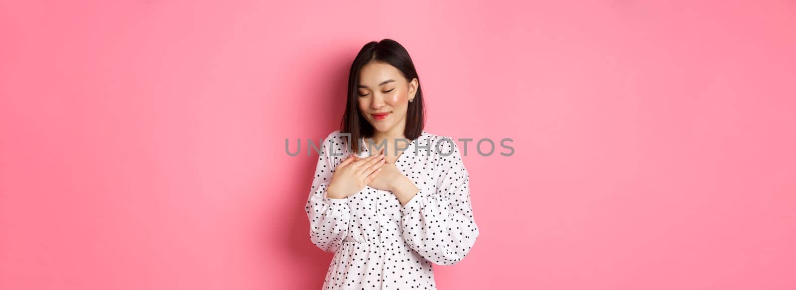 Beautiful korean girl in trendy spring dress dreaming, holding hands on heart, smiling with closed eyes, imaging something or having heartwarming memory, pink background.