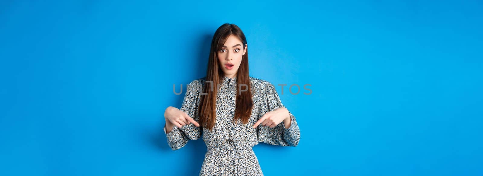 Excited young woman in dress pointing fingers down at logo, looking intrigued at camera, standing on blue background by Benzoix
