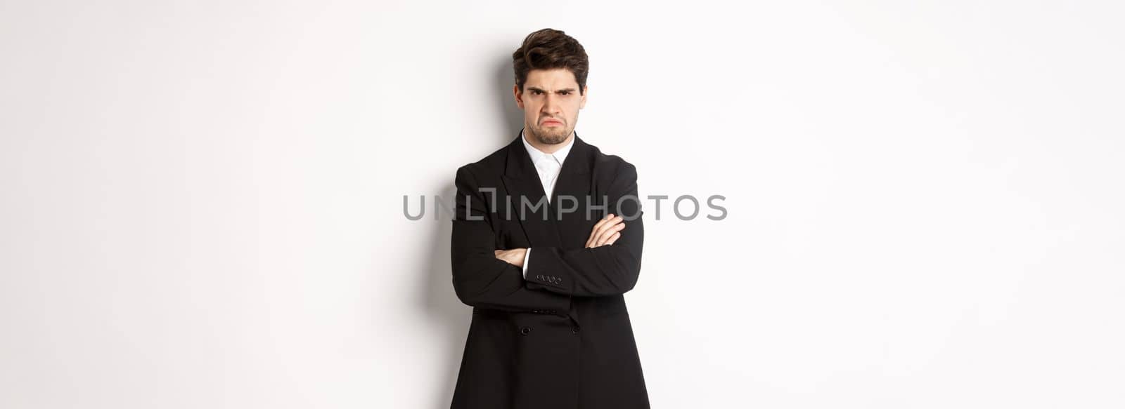 Portrait of angry handsome man in black suit, cross arms on chest and looking offended, frowning and pouting, being mad at someone, standing over white background.