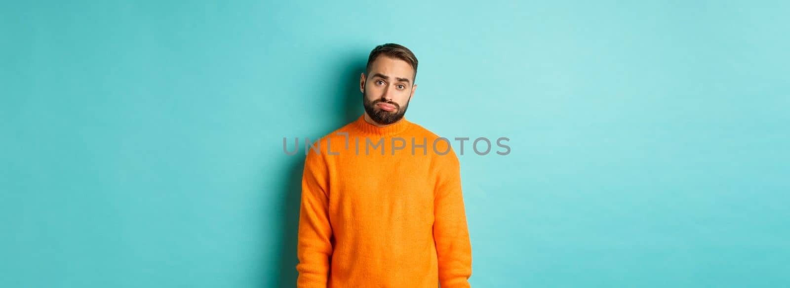 Sad man looking at camera, sulking and feeling distressed, standing over light blue background.