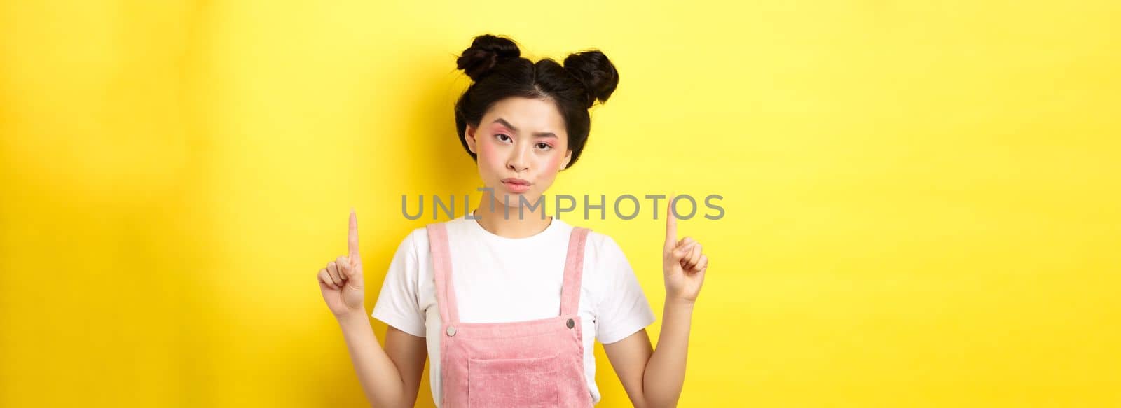 Annoyed and tired asian girl with pink glamour makeup, pointing fingers up and look bothered, standing on yellow background.