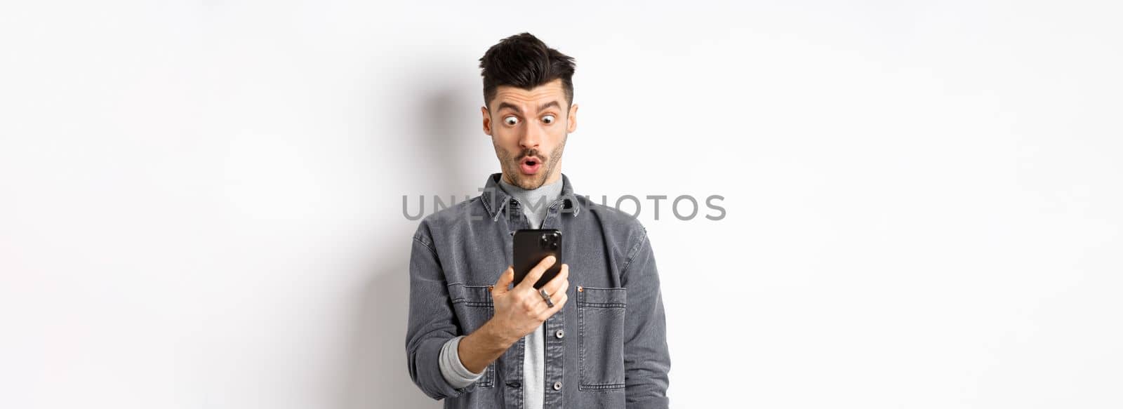 Man look surprised at smartphone screen, receive exciting news on phone, standing against white background by Benzoix