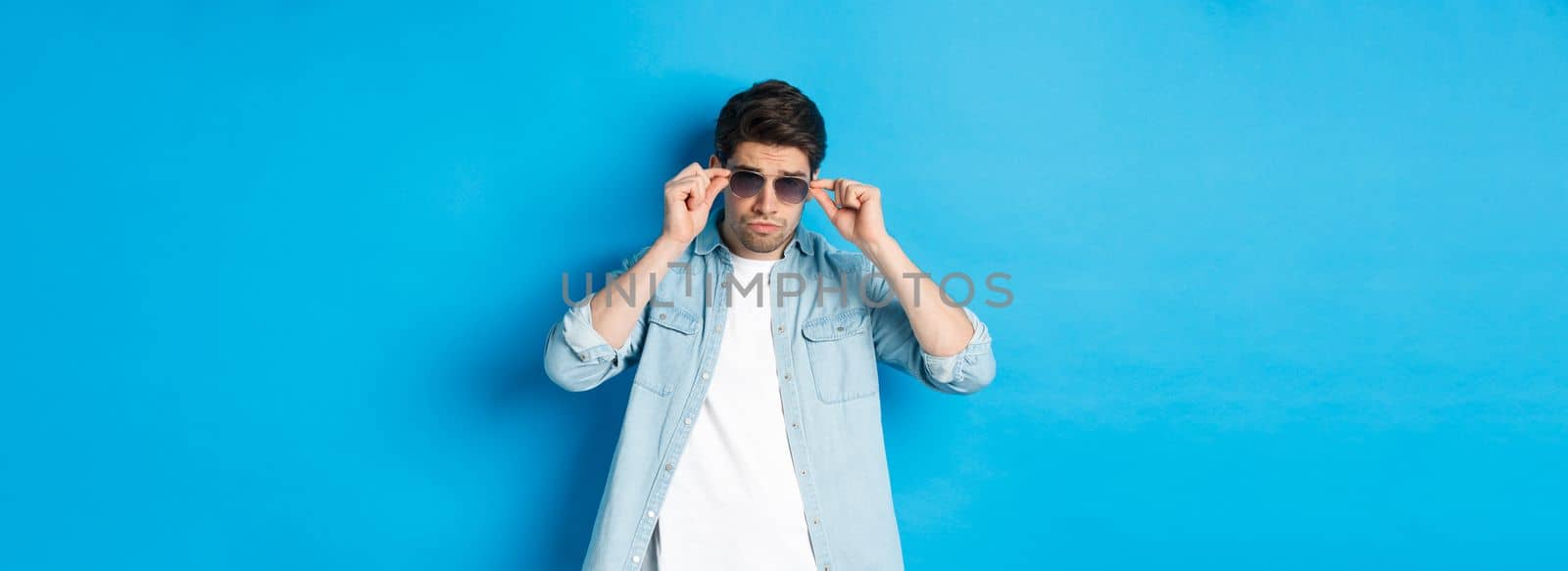 Confident macho man put on sunglasses, looking cool and sassy, standing over blue background.