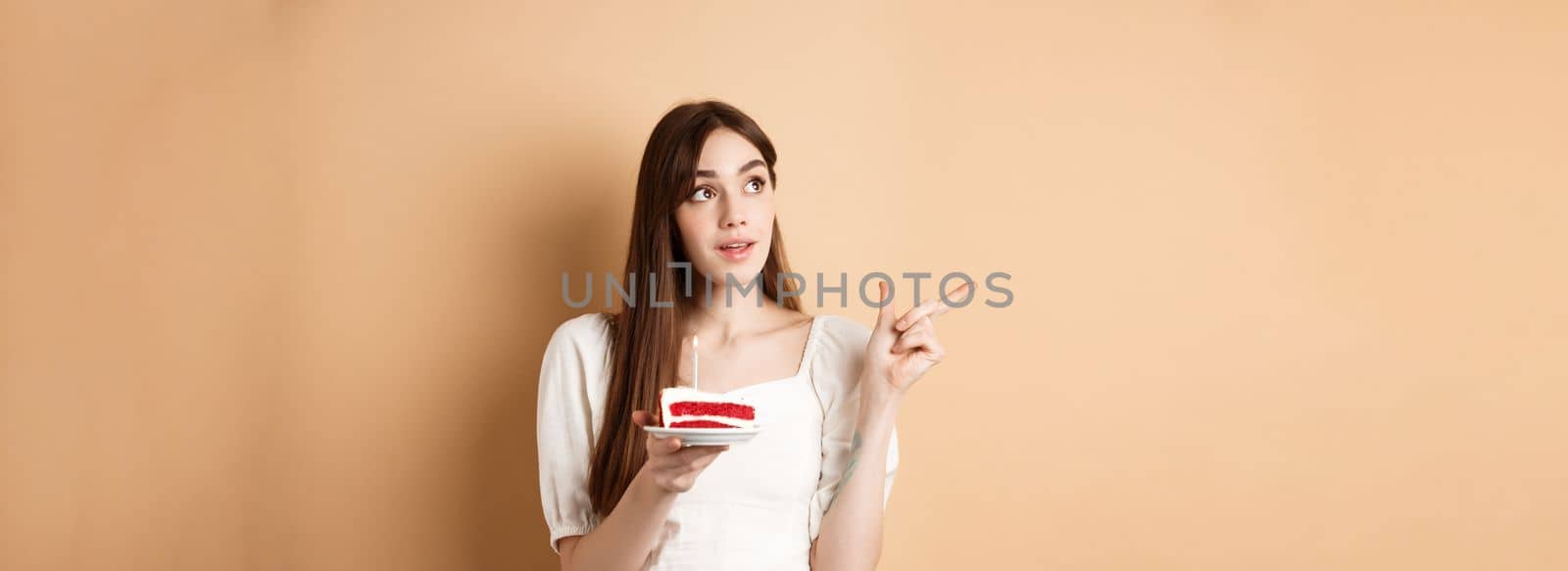 Thoughtful birthday girl holding cake with candle, thinking of wish and pointing left at logo, standing on beige background.