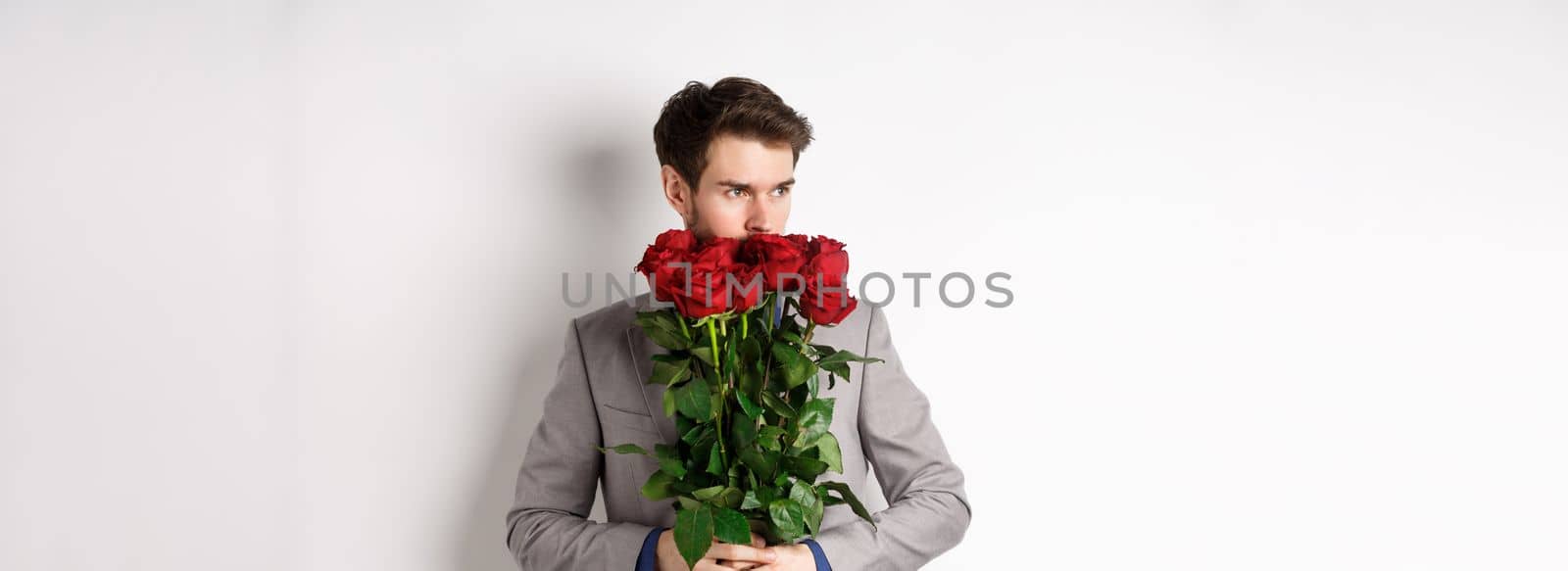 Romantic man in suit smell bouquet of roses and looking pensive, standing over white background. Concept of valentines day.