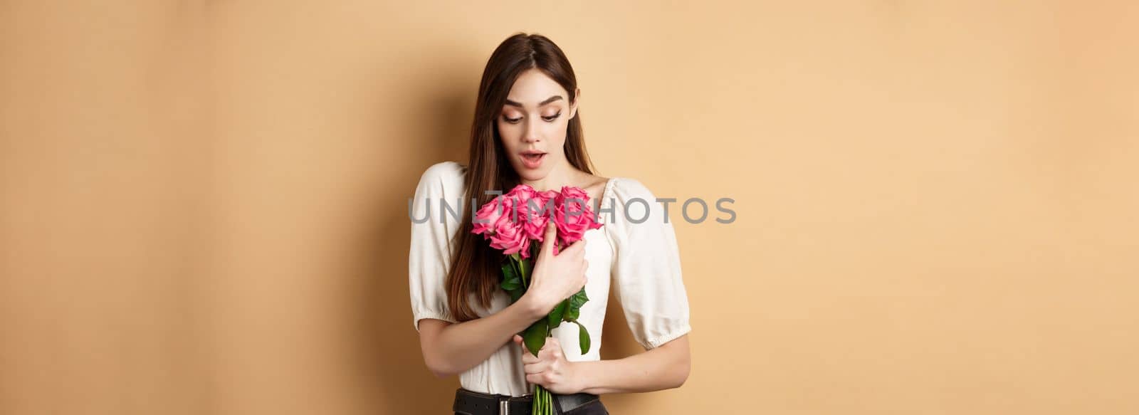 Valentines day. Surprised girl receive gift from lover on date, looking amazed at beautiful bouquet of flowers, holding roses on beige background by Benzoix
