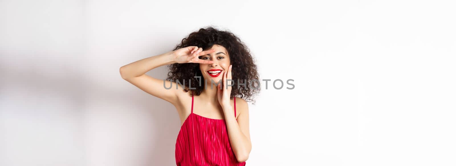 Beauty and fashion. Romantic woman with curly hair and red dress, showing v-sign and smiling happy at camera, standing on white background.