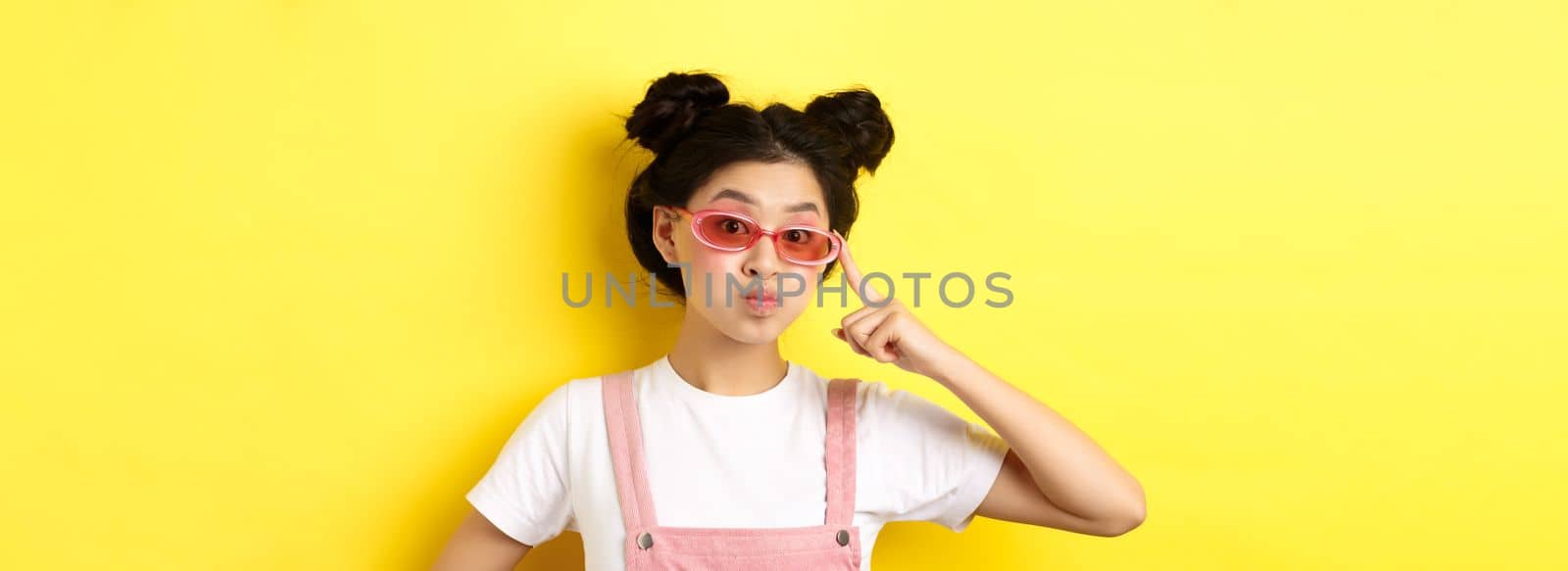 Stylish teen asian girl wearing sunglasses and pink overalls, standing on yellow background.