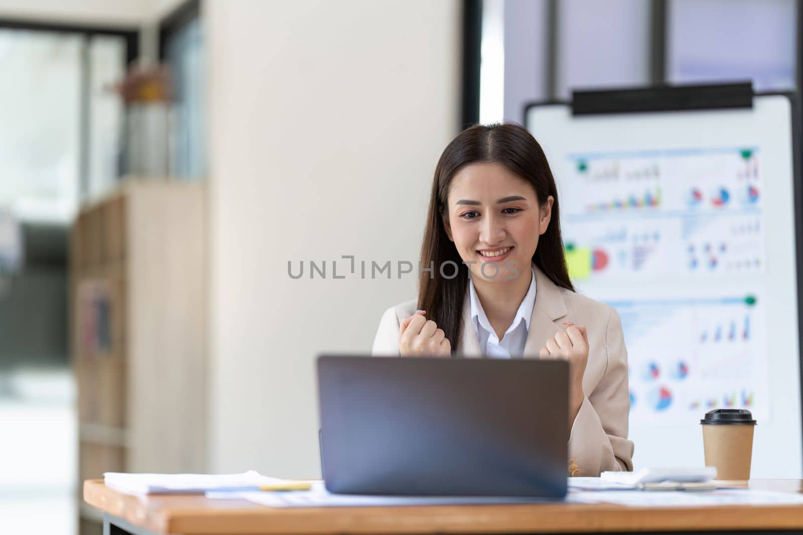 Successful young Asian business woman achieving goals excited raised hands rejoicing with laptop in the office. New startup project concept by nateemee
