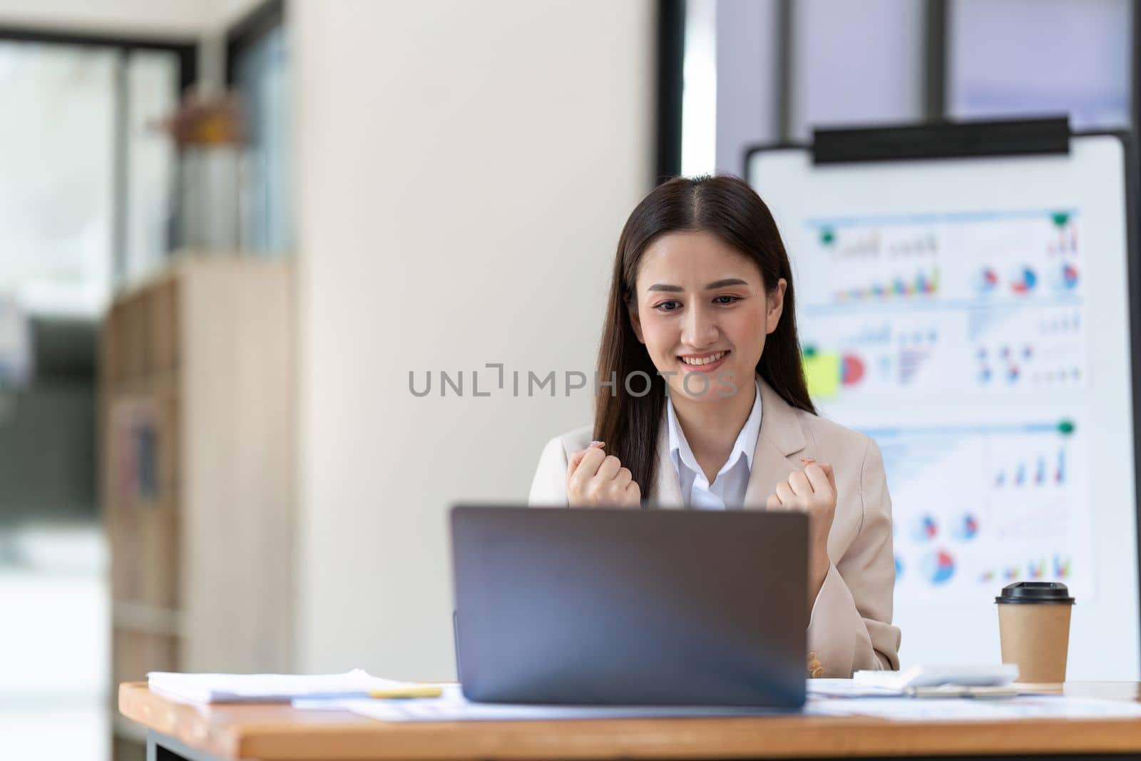 Successful young Asian business woman achieving goals excited raised hands rejoicing with laptop in the office. New startup project concept.