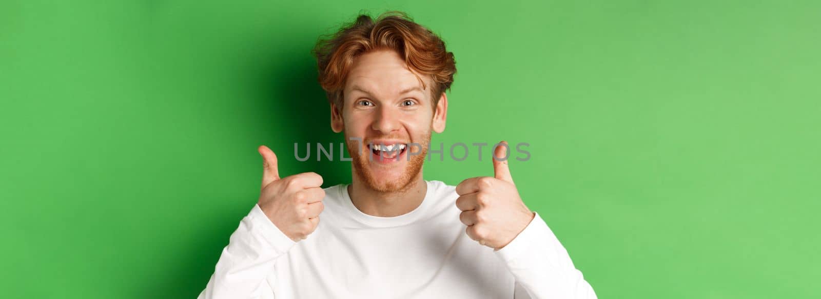 Emotions and fashion concept. Close up of cheerful redhead man showing thumbs-up and say yes, approve and like something cool, green background.