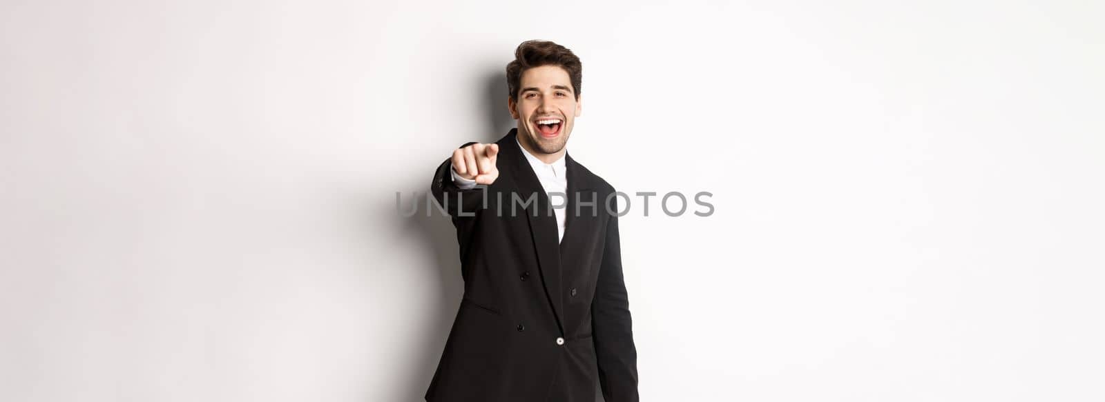 Concept of new year party, celebration and lifestyle. Portrait of handsome stylish man in black suit, smiling and pointing finger at camera, standing over white background.