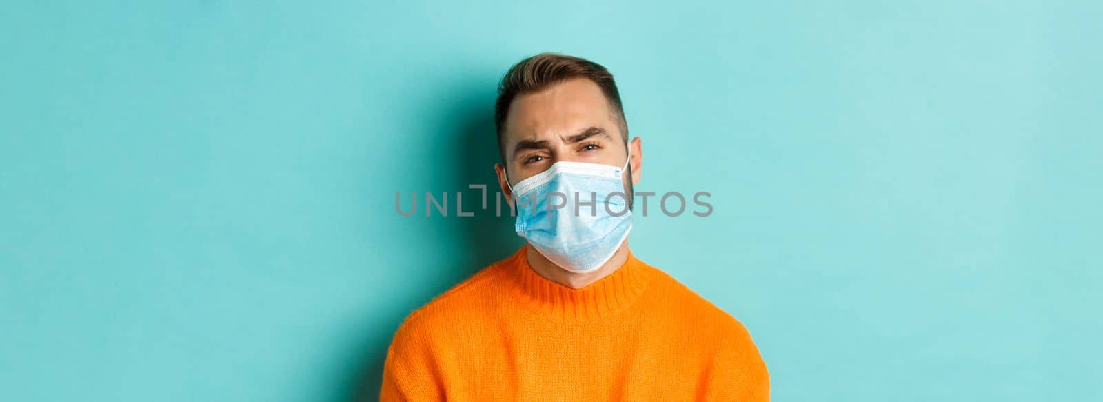 Covid-19, social distancing and quarantine concept. Upset man in face mask complaining, looking displeased and tired, standing against light blue background by Benzoix