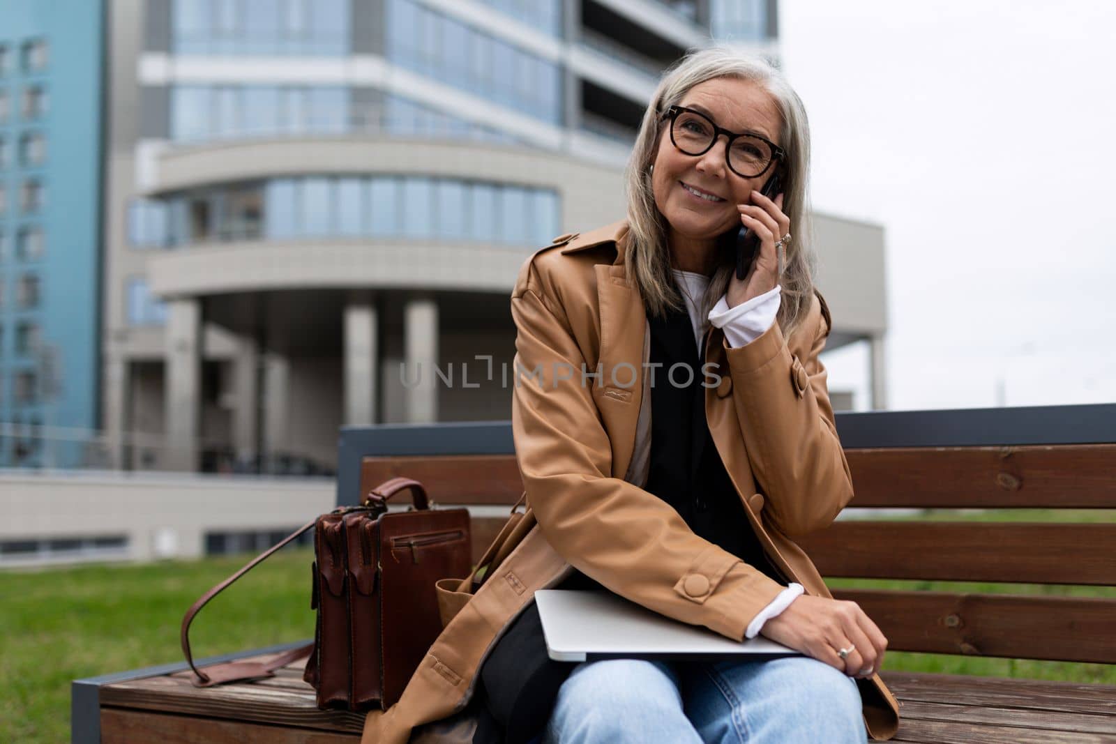 mature woman talking on the phone while sitting on a bench in an office building outside.