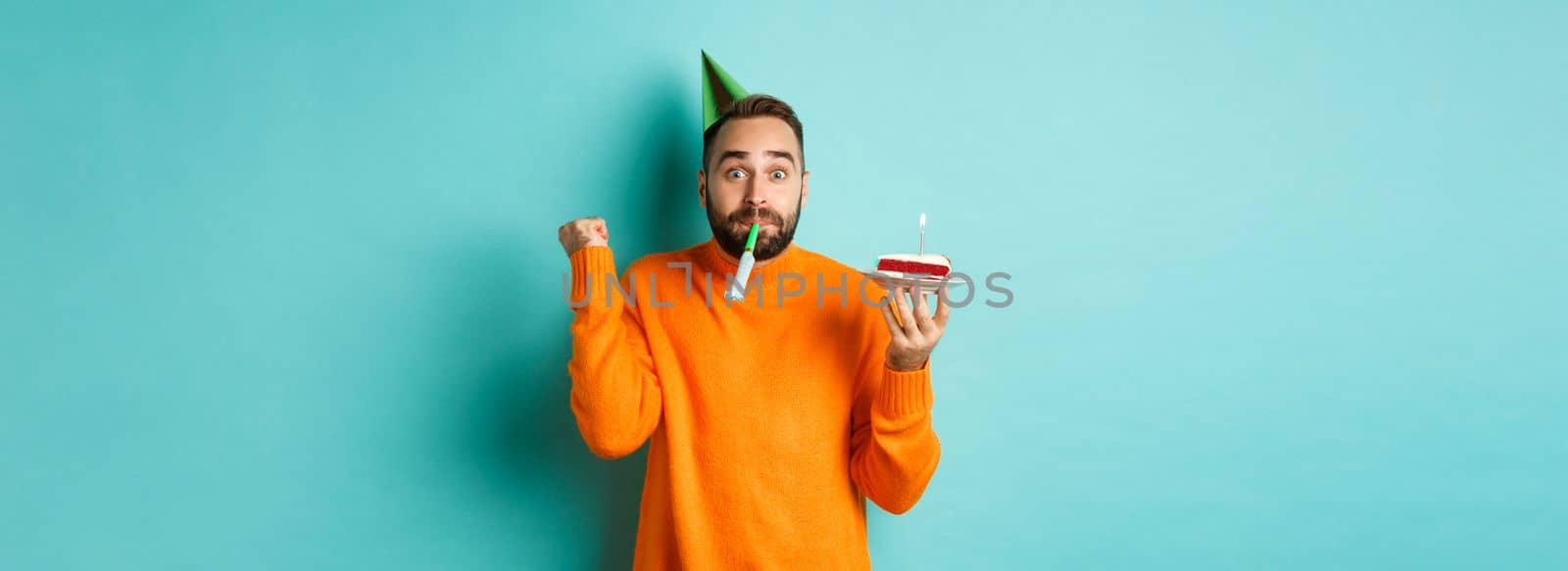 Happy birthday guy celebrating, wearing party hat, blowing wistle and holding bday cake and doing fist pump excited, standing over turquoise background.