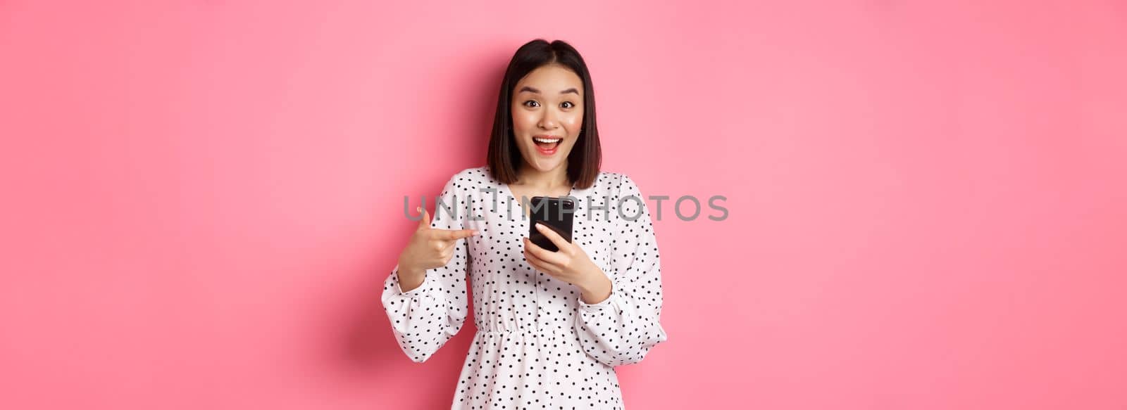 Online shopping and beauty concept. Amazed and happy asian woman pointing at mobile phone, talking about internet promo offer or app, standing over pink background.