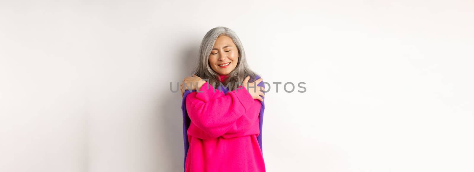 Valentines day and holidays concept. Lovely asian senior woman in pink sweater hugging herself with eyes closed, smiling, standing over white background.