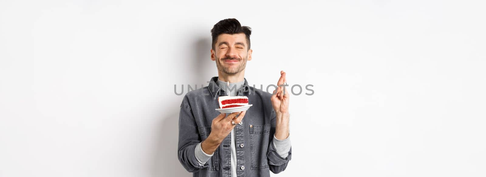 Hopeful birthday guy making wish with fingers crossed, holding bday cake on party, standing against white background by Benzoix