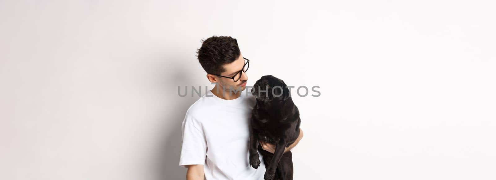 Handsome young man holding his black pug, dot and pet owner looking each other faces, standing against white background.