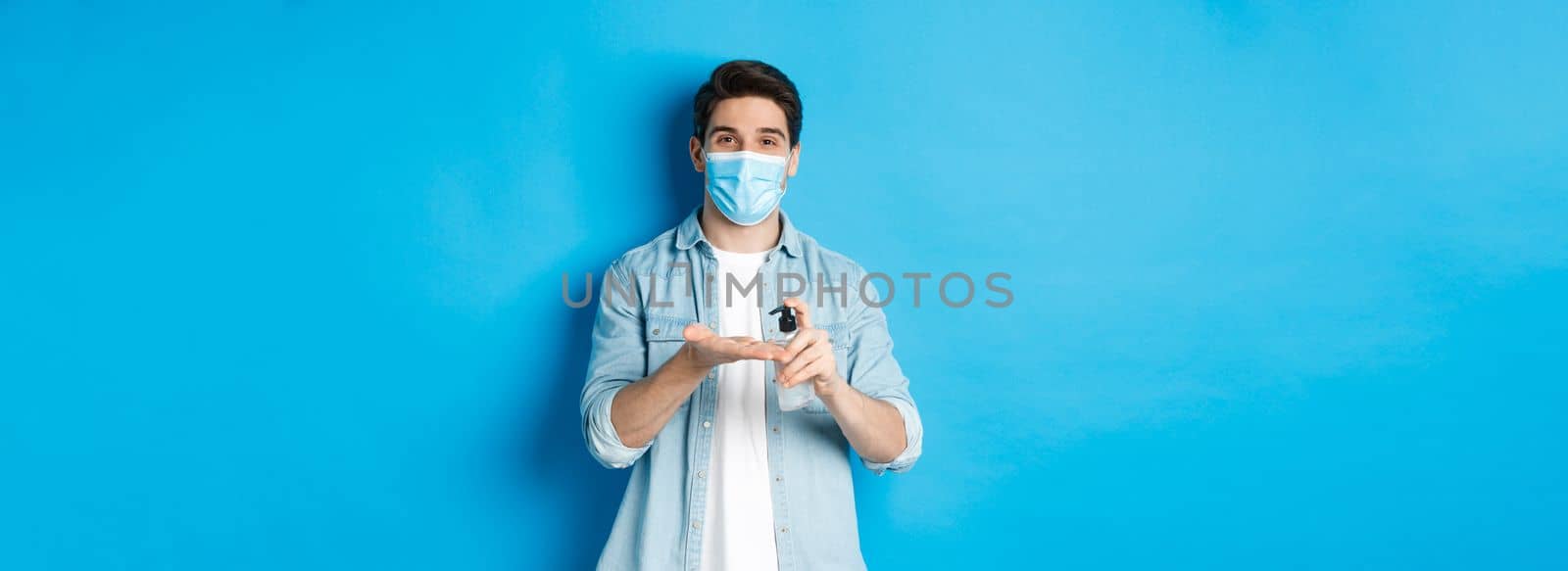 Concept of covid-19, pandemic and social distancing. Handsome young man in medical mask disinfecting hands with sanitizer, using antiseptic, standing against blue background by Benzoix