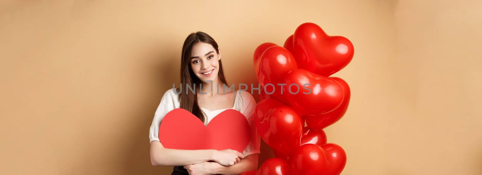 Valentines day and love concept. Lovely girl hugging big romantic heart cutout and smiling, standing near red balloons on beige background by Benzoix