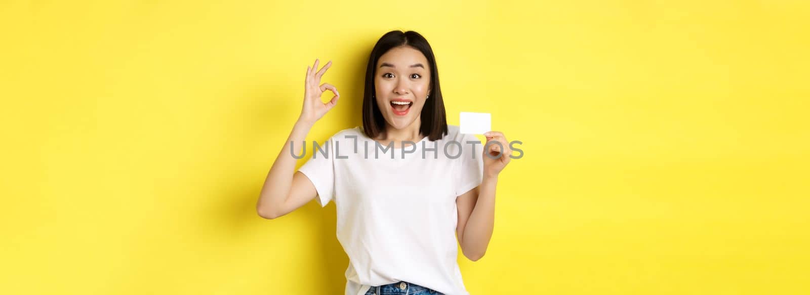 Young asian woman in casual white t-shirt showing plastic credit card and okay gesture, recommend bank, smiling at camera, yellow background.