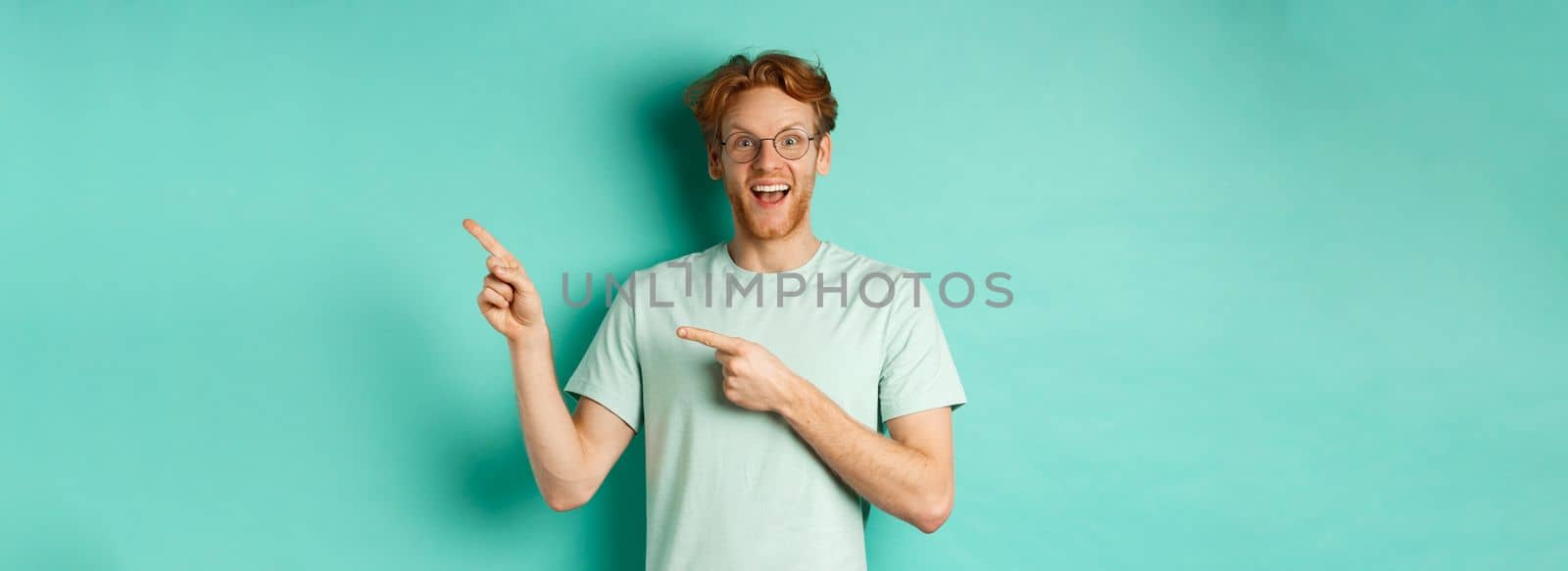 Handsome smiling man with red hair and beard, looking amused and pointing at upper right corner, showing promo offer, standing over mint background by Benzoix