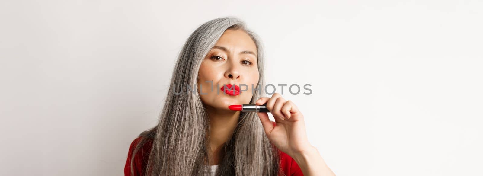 Beauty and makeup concept. Beautiful asian elderly woman pucker lips, showing red lipstick and looking sassy at camera, white background.