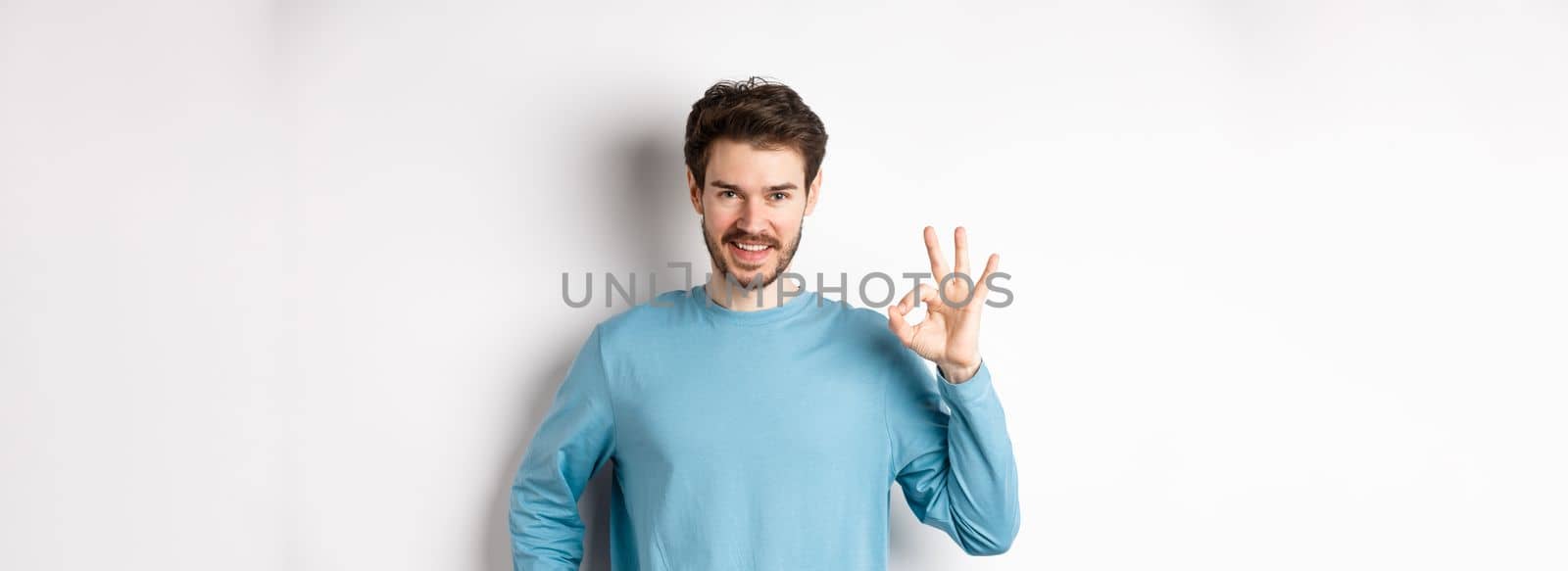 Okay. Handsome smiling man showing ok sign and looking confident, recommend or praise something good, standing over white background by Benzoix