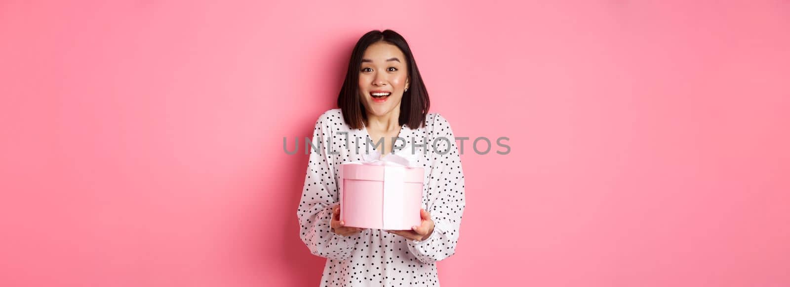Surprised and happy asian woman receiving cute box with gift, standing amazed over pink background.