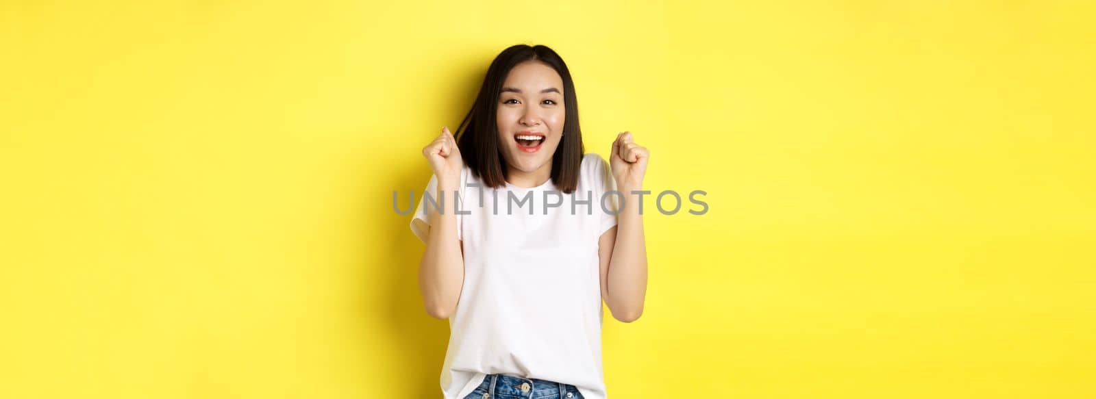 Carefree asian girl dancing and having fun, posing in white t-shirt against yellow background.