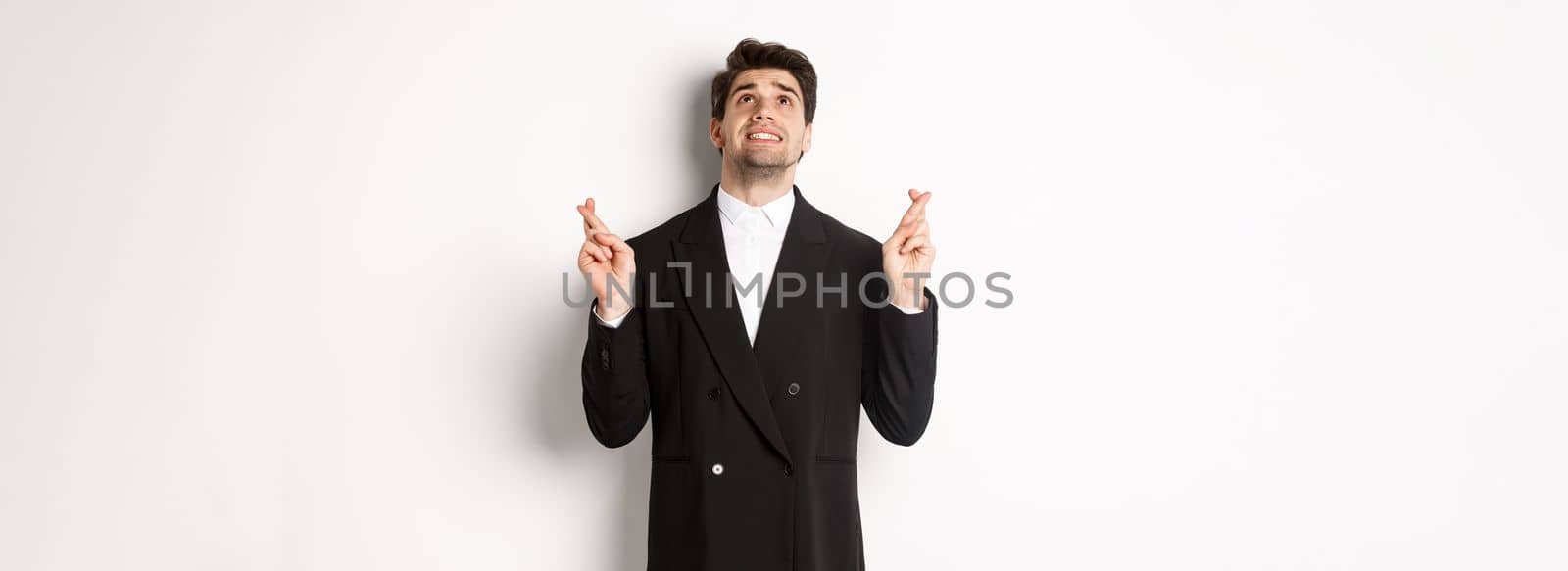 Portrait of tensed and worried handsome businessman, crossing fingers and looking up, begging god, making a wish, standing against white background in black suit.