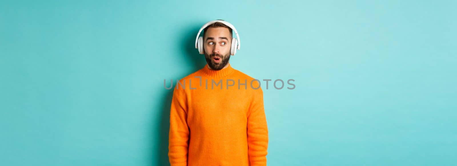 Amazed adult man listening music in headphones, looking at camera impressed with sound, standing over turquoise background.