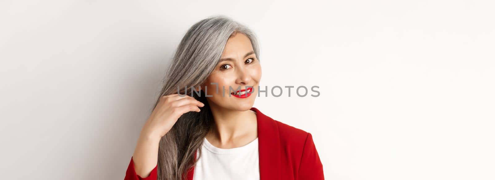 Beauty and haircare concept. Close up of elegant asian senior woman showing shiny and healthy grey hair, smiling and looking aside, white background.