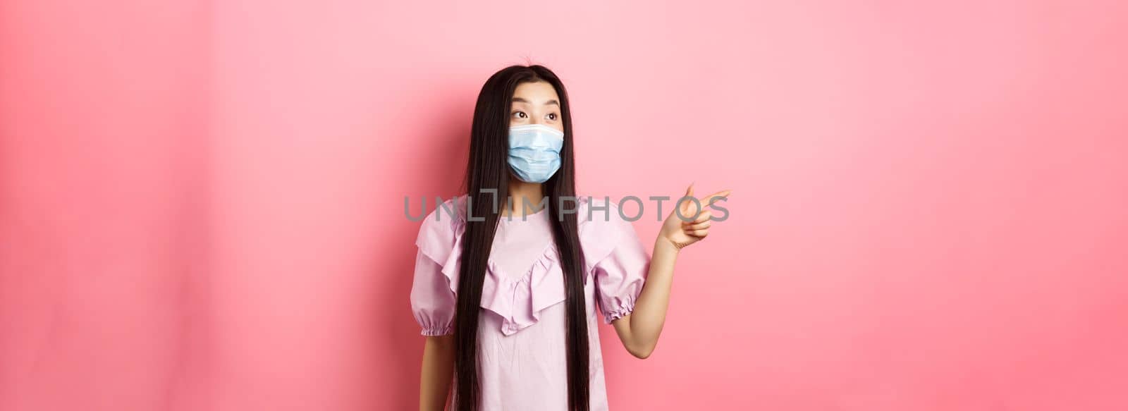 Coronavirus, quarantine and lifestyle concept. Excited asian teen girl in face mask pointing, looking left at logo with amazed face, checking out promotion, white background.