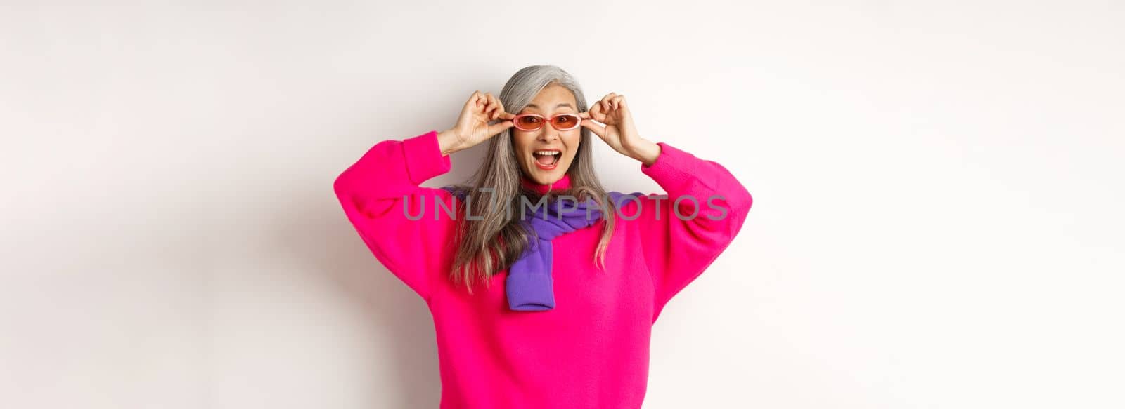 Fashionable asian senior woman trying new sunglasses, looking amazed and cheerful at camera, standing in trendy sweater against white background.