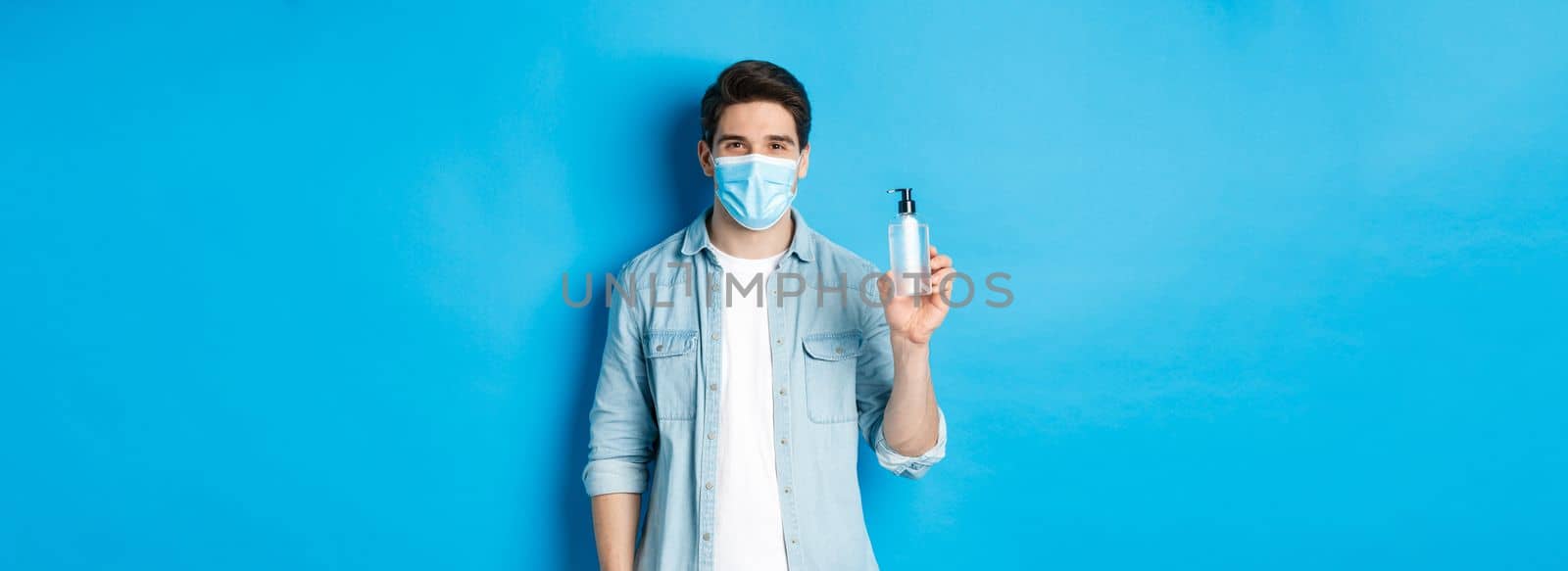 Concept of covid-19, pandemic and social distancing. Young man in medical mask showing hand sanitizer, standing against blue background by Benzoix