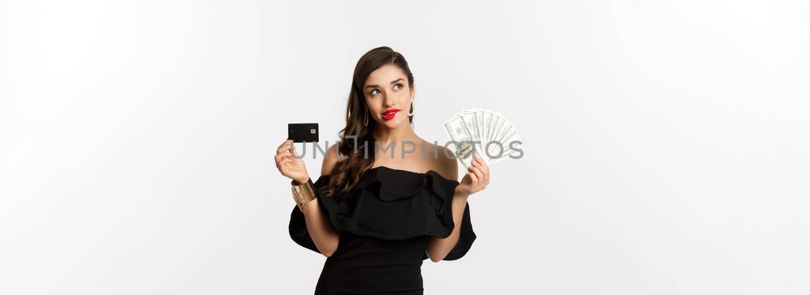 Fashion and shopping concept. Elegant woman in black dress, red lipstick, holding money and credit card, making choice, thinking, standing over white background.