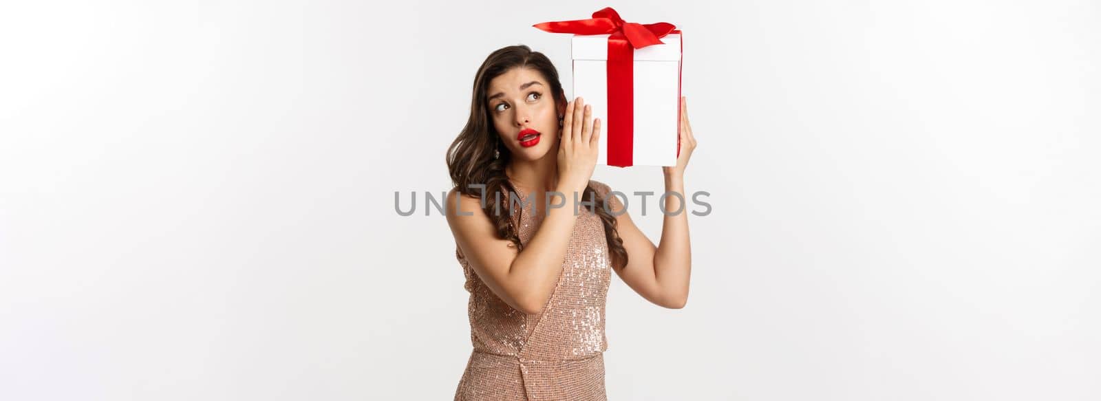 Holidays, celebration concept. Beautiful woman in luxury dress trying to guess what inside gift box, shaking Christmas present with curious face, white background by Benzoix