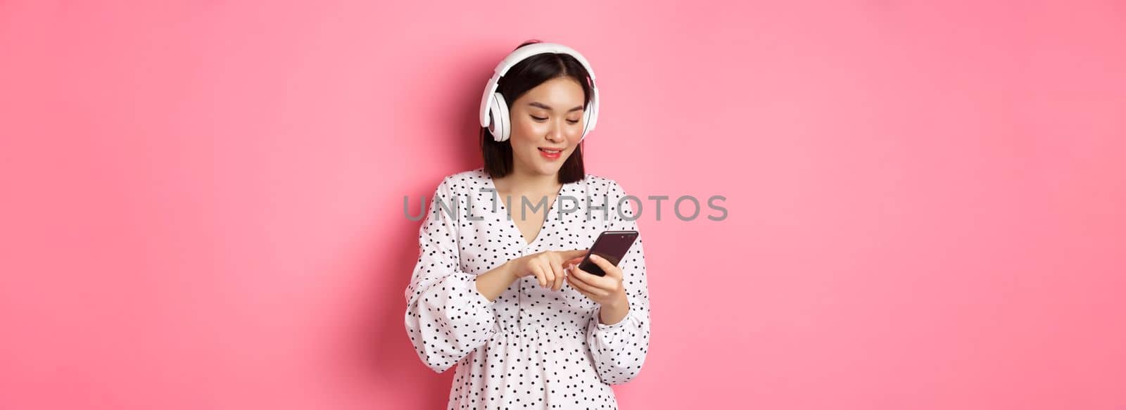 Beautiful asian woman texting message on smartphone, listening music in headphones, standing over pink background.