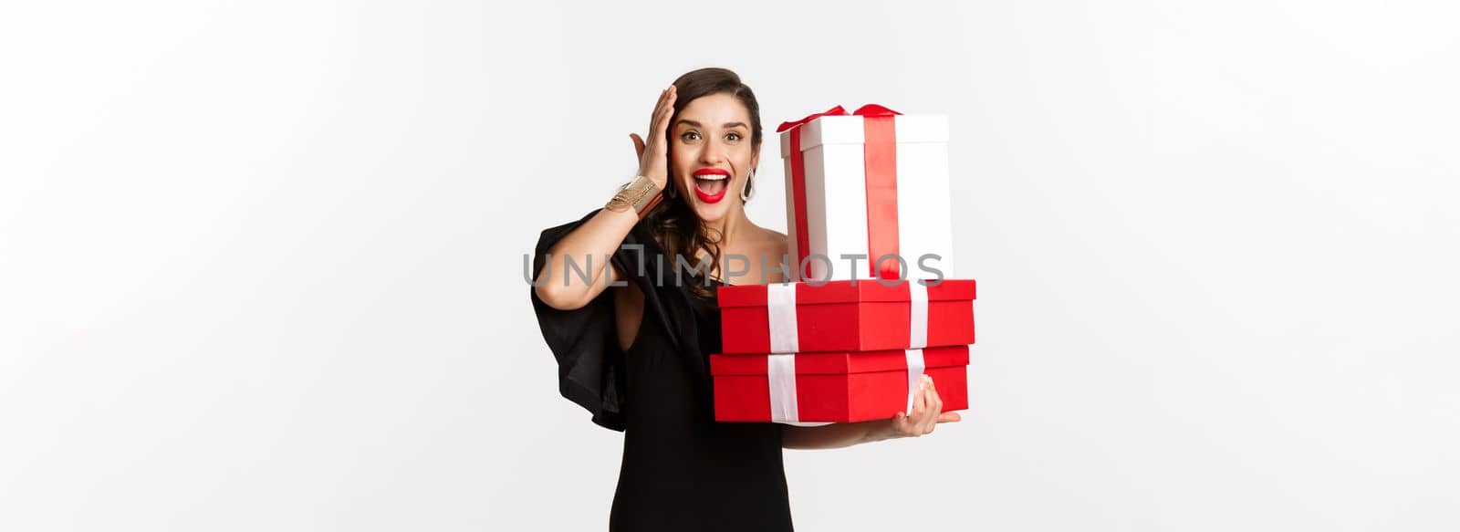 Celebration and christmas holidays concept. Excited and happy woman receive gifts, holding xmas presents and rejoicing, standing in black dress over white background by Benzoix