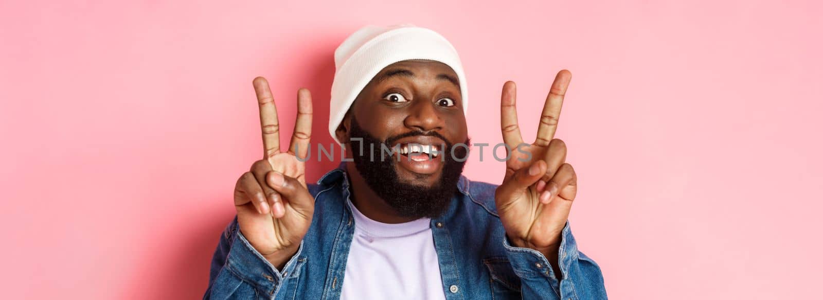 Close-up of cheerful african-american hipster in beanie smiling, showing peace signs, standing happy against pink background by Benzoix