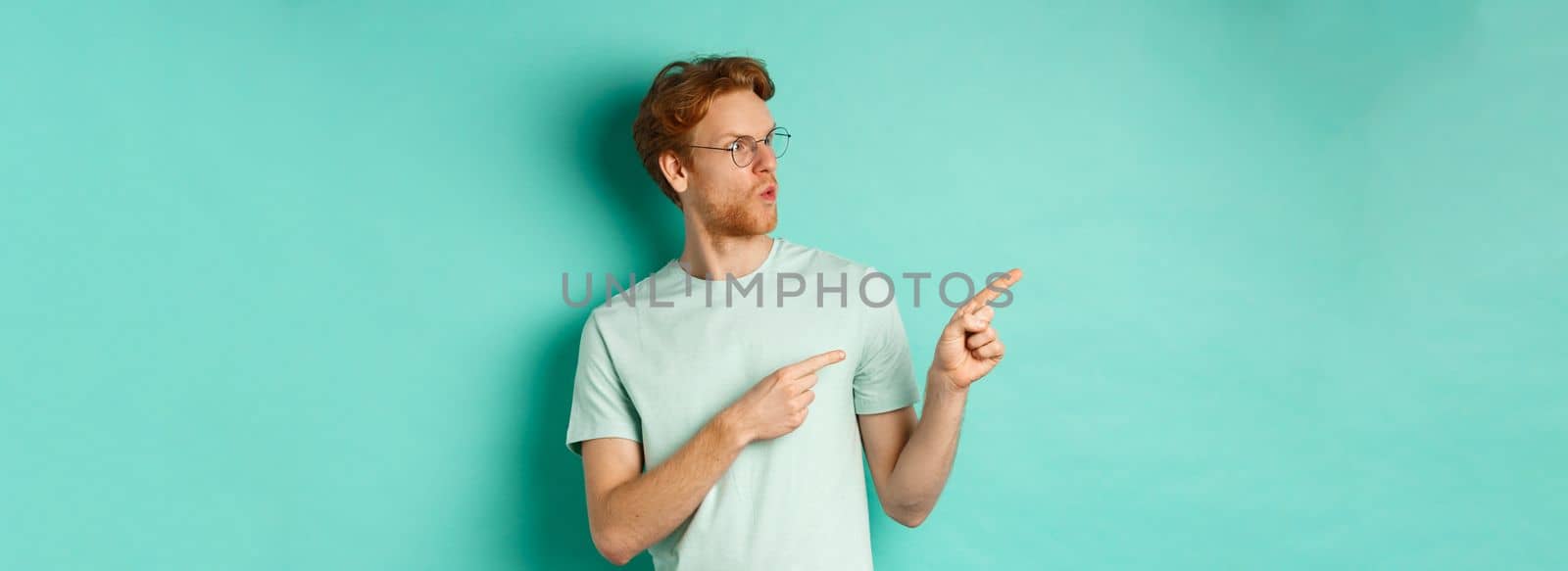 Impressed redhead man in glasses and t-shirt, pointing fingers and looking left at promo offer, staring happy, standing over turquoise background.