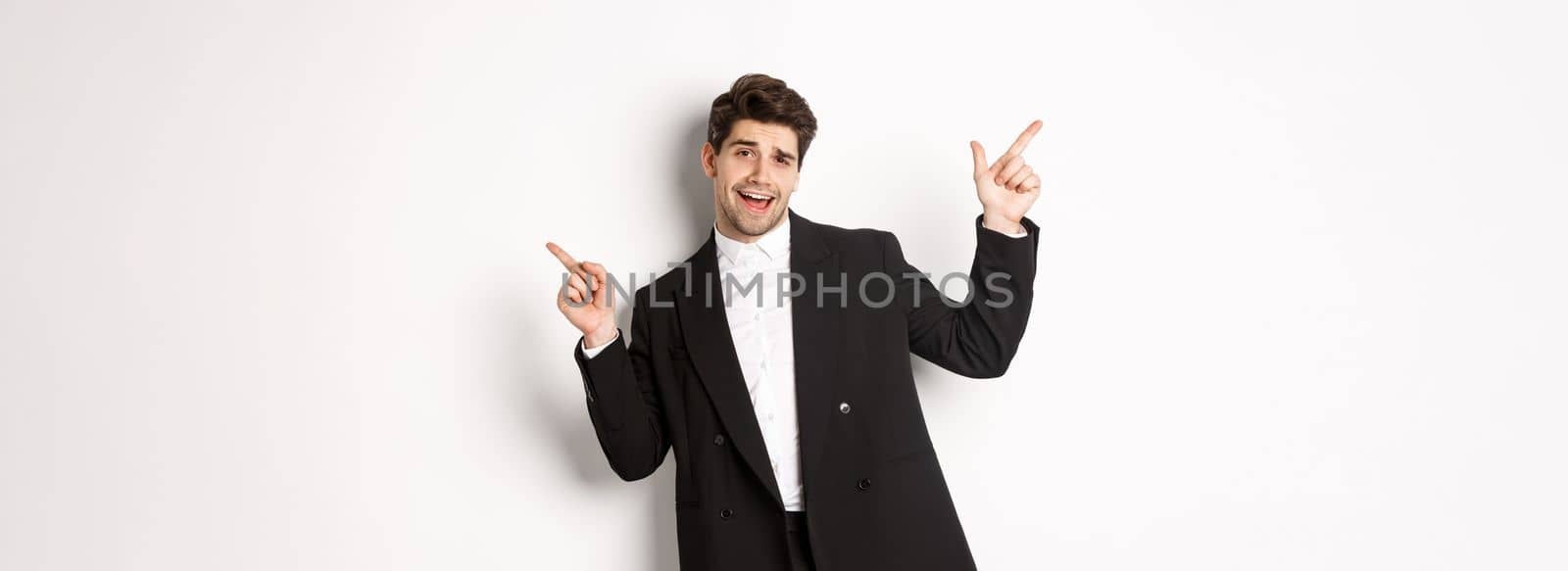Handsome guy dancing and partying in black suit, pointing fingers sideways, showing two promo banners, standing against white background.